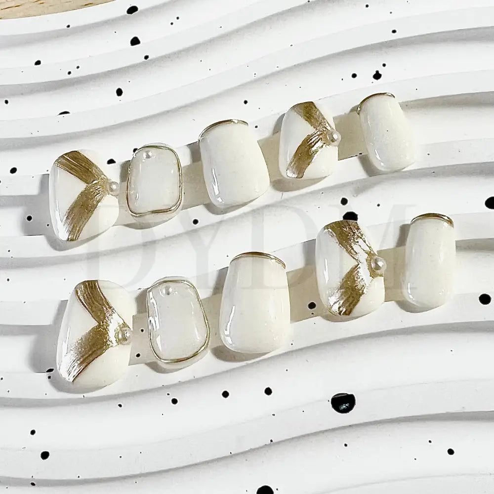 Golden Ivory handmade press on nails shows off-white ivory base color enhanced by intricate golden details. The ring finger and thumb showcase captivating golden chrome ribbon accents, while the remaining nails feature a delicate thin golden line French tip